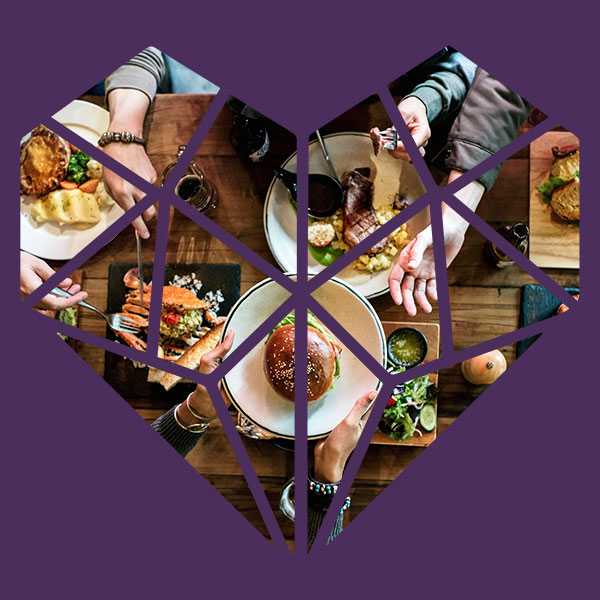 An overhead view of a long wood dinner table with food that has fresh ingredients on it and the hands of friends reaching for the food